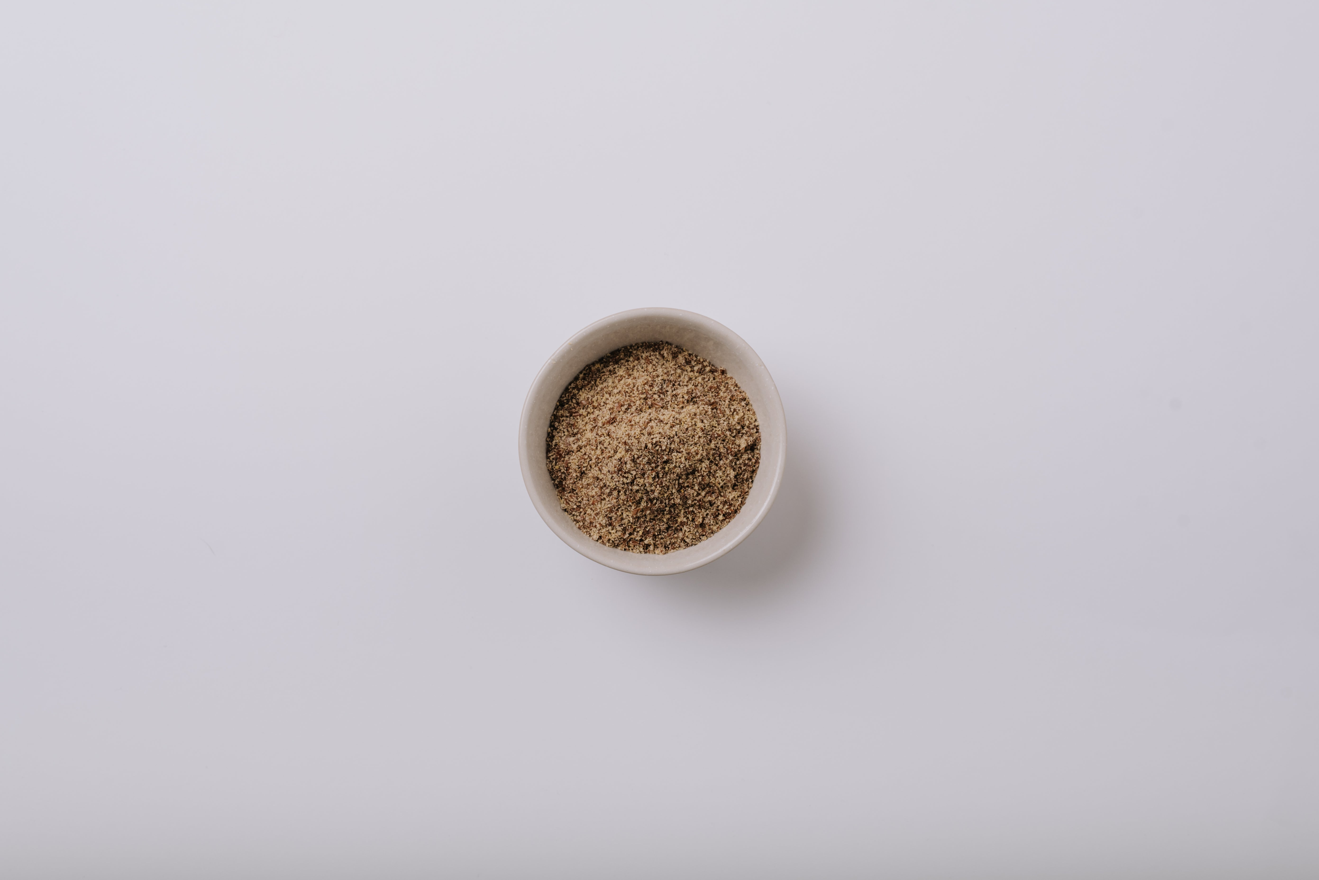 Small white bowl filled with ground flaxseed on a light grey background