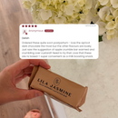 A hand holds a wrapped Lila Jasmine milk support lactation bar, with a review card overlaid on the image and white flowers in the background.