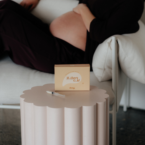 Box labeled 'Mother's Gold Colostrum Collection Kit' on a small table with a blurred pregnant person sitting in the background.