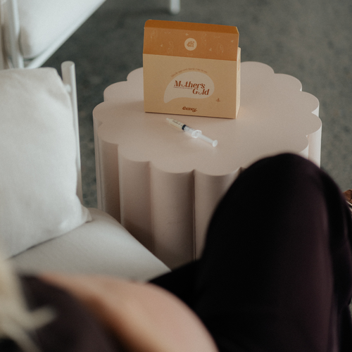 Box labeled 'Mother's Gold Colostrum Collection Kit' on a small table with a syringe, pregnant woman sitting on a couch.
